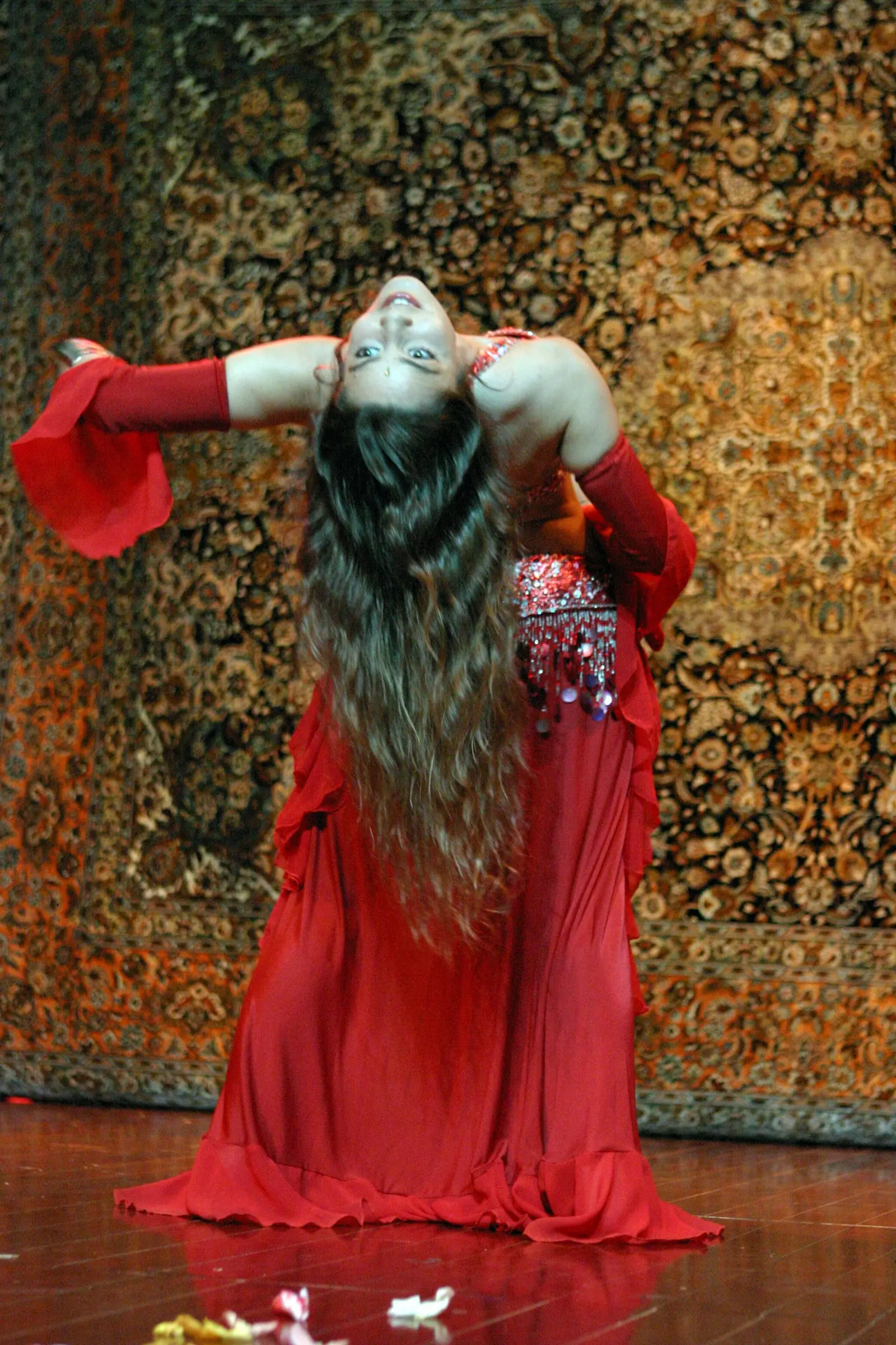 Ana in a deep backbend on stage, in a red costume with her long hair falling, before an oriental carpet