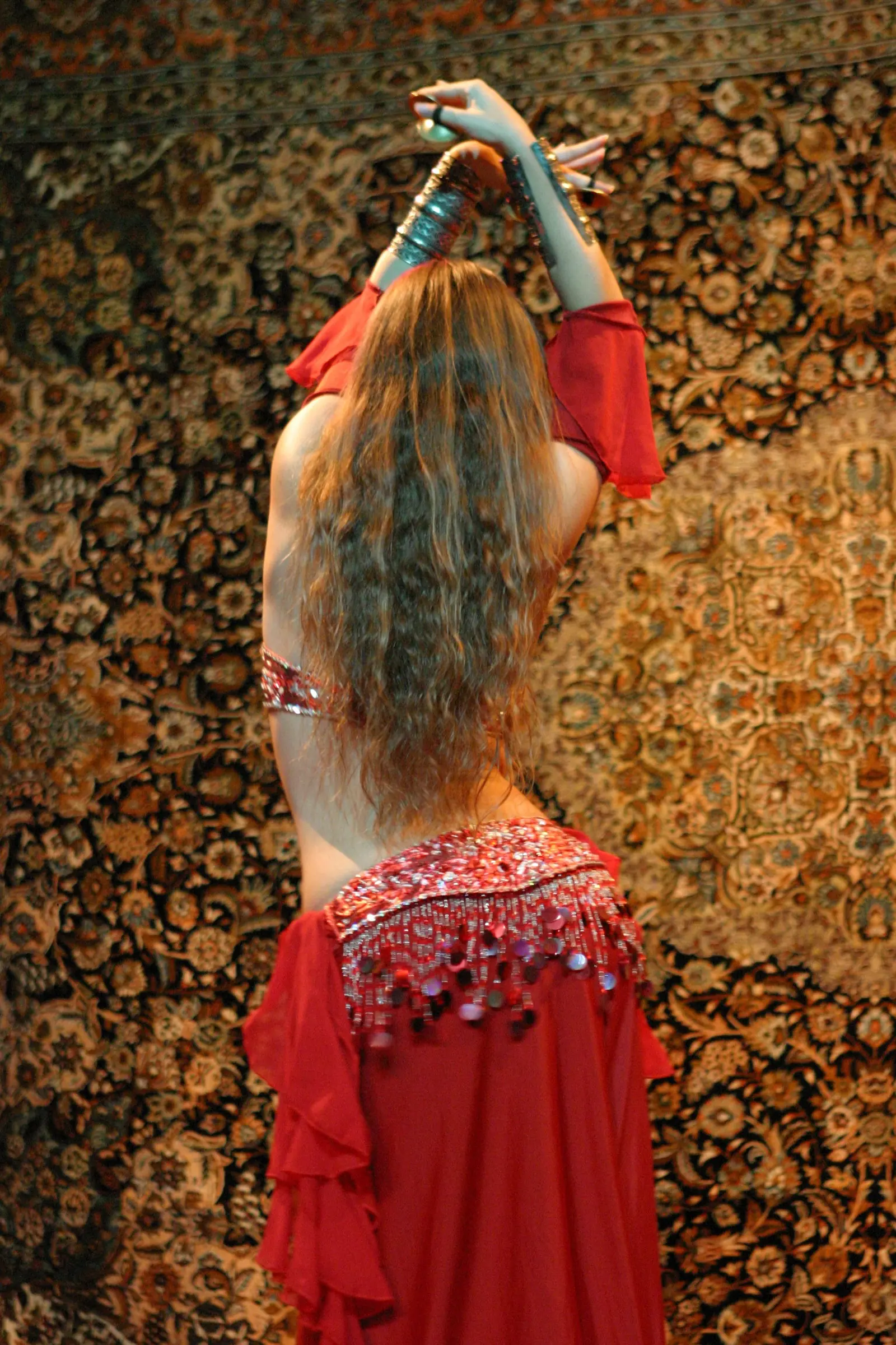Ana seen from behind, with long wavy hair and a red costume, arms raised before an oriental carpet
