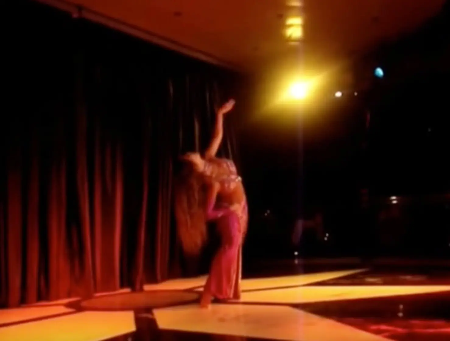 Ana dancing alone on a dark stage under a spotlight, with red curtains behind her