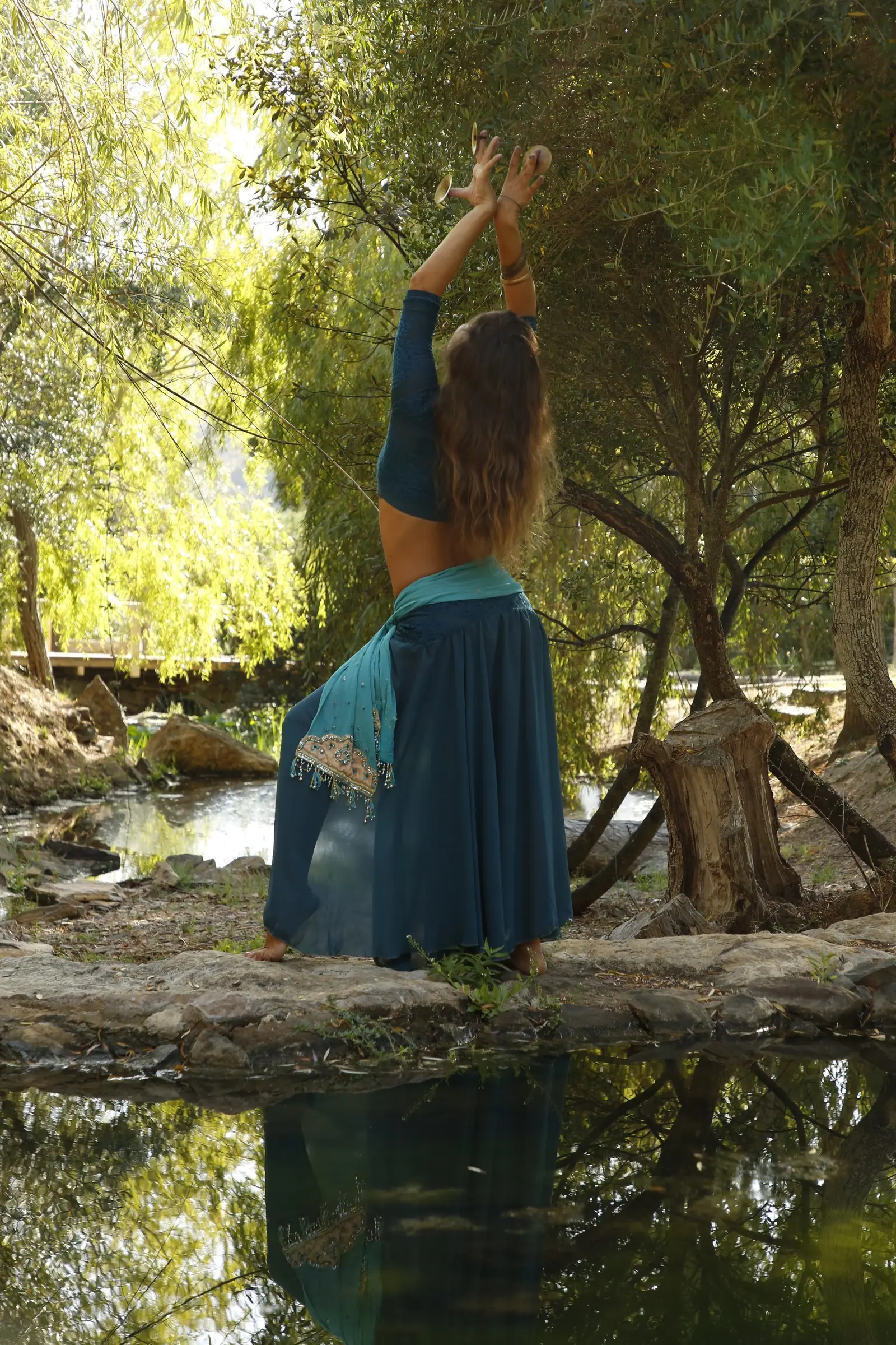 Ana seen from behind, arms raised with finger cymbals, by a stream among trees