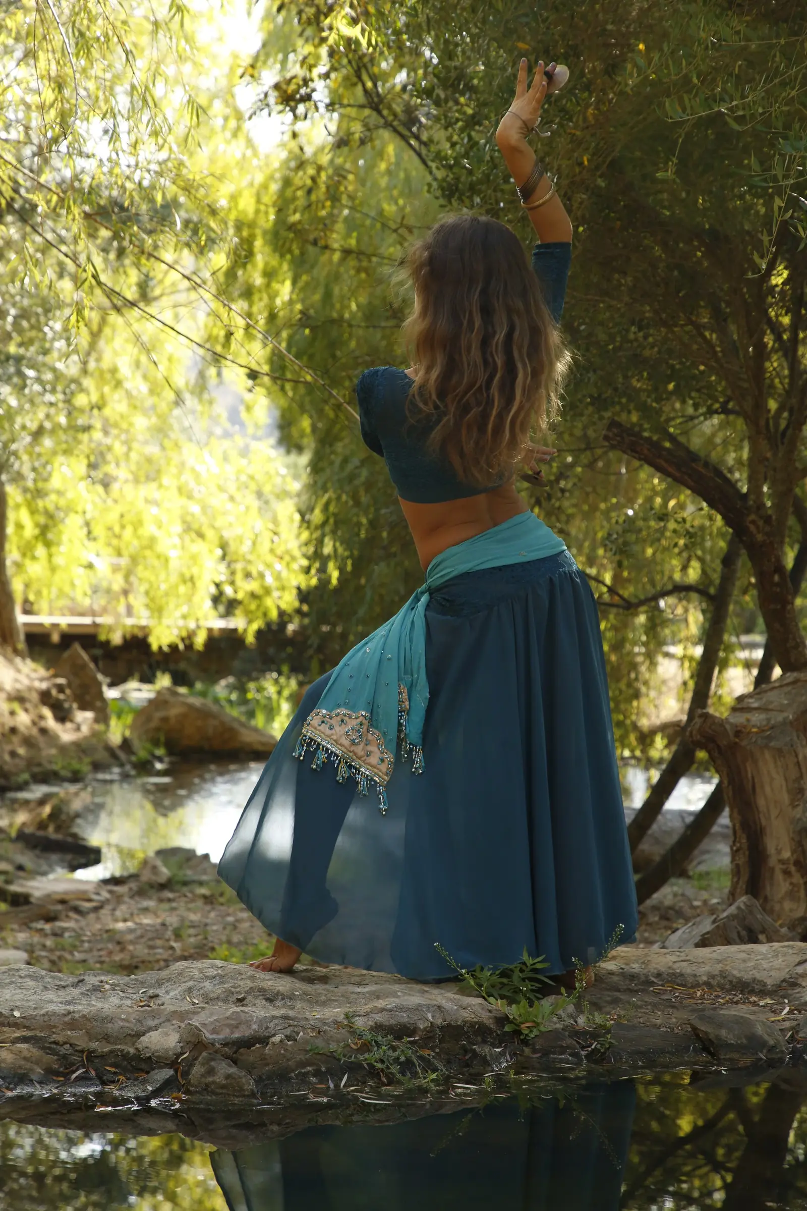 Ana seen from behind on a rock by a stream, one arm raised with finger cymbals