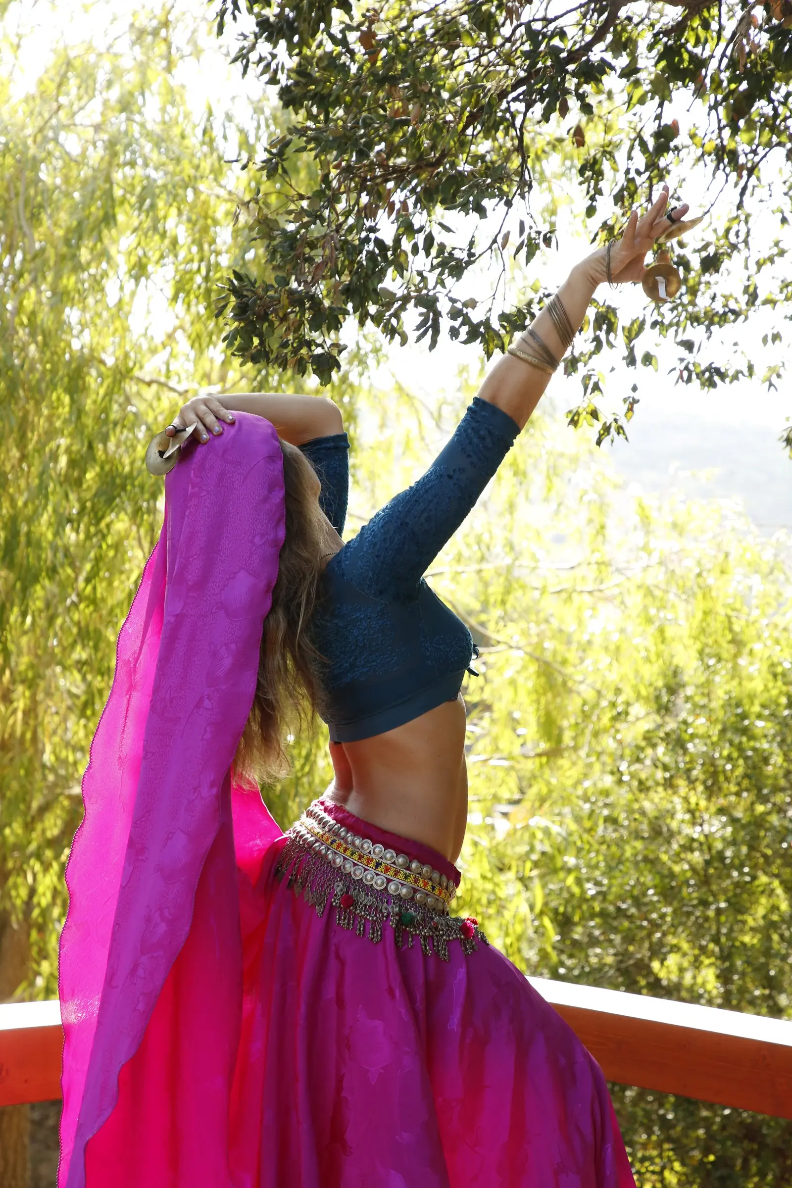 Ana stretches one arm up with finger cymbals and a fuchsia veil, by a wooden rail
